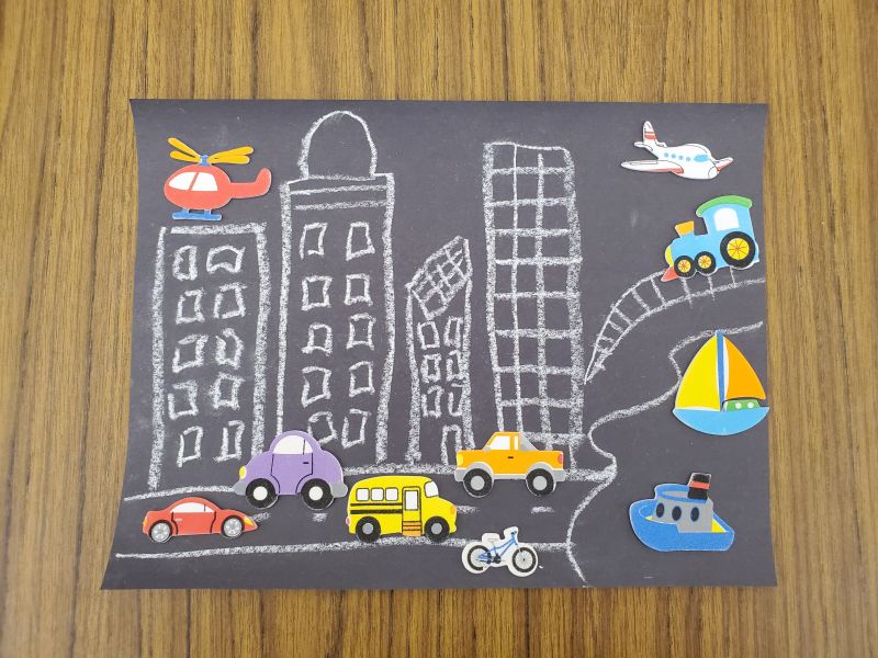 Photo of craft: white chalk drawing on black paper of buildings, a road, a harbor, and a railroad, with colorful vehicle stickers such as cars, buses, boats, trains, and helicopters.