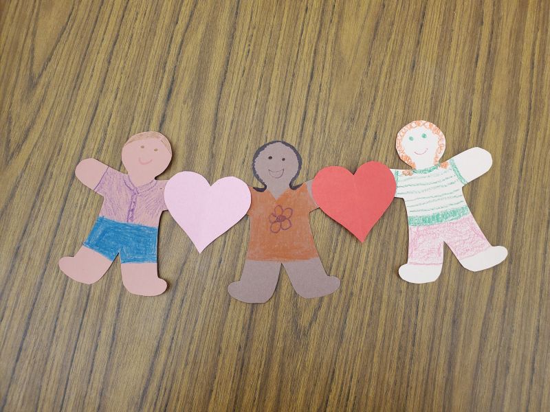 Photo of three generic construction paper people figures of different skin tones, each colored with unique clothes and hair, connected at the arms with two hearts.