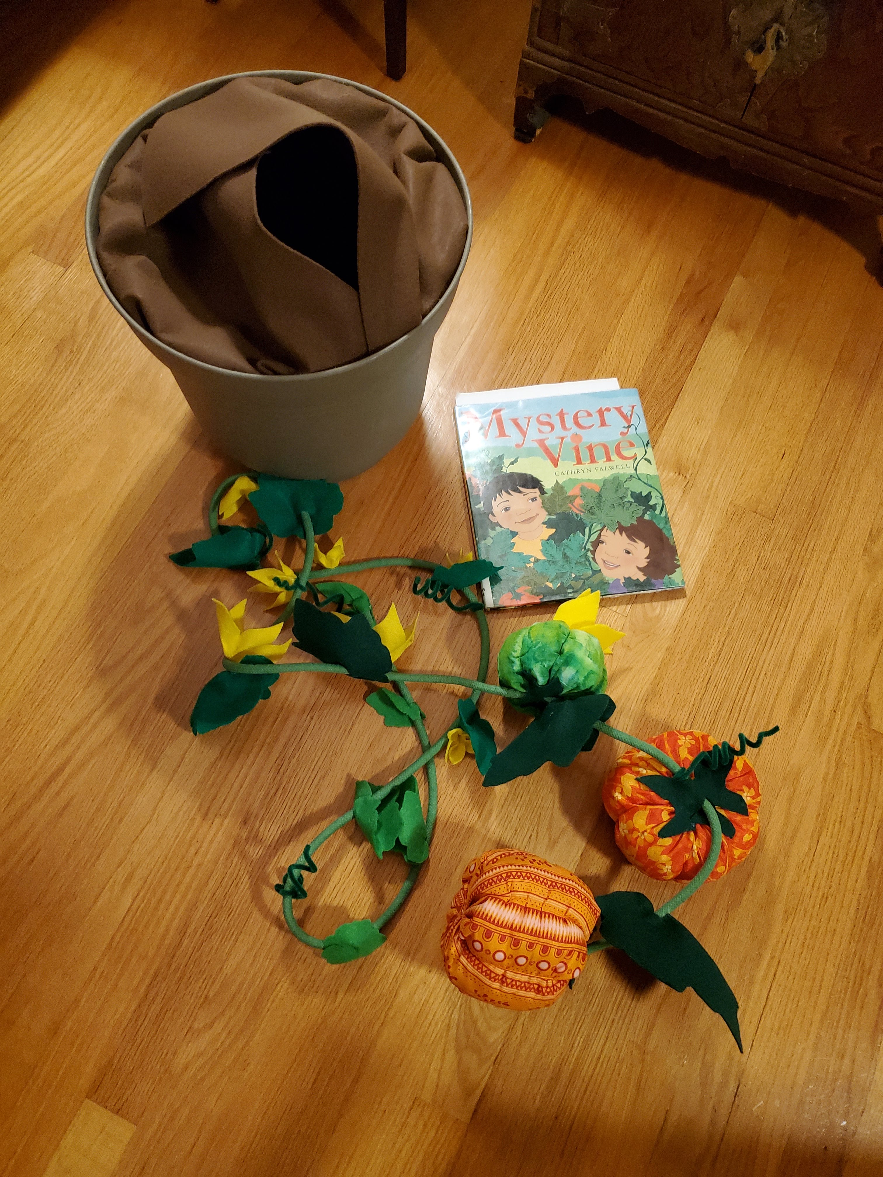 photo of mystery vine prop extended - a big plastic pot with brown felt, and a green vine with felt leaves, flowers, pipe cleaner vines, a green pumpkin, and two orange pumpkins made of fabric.