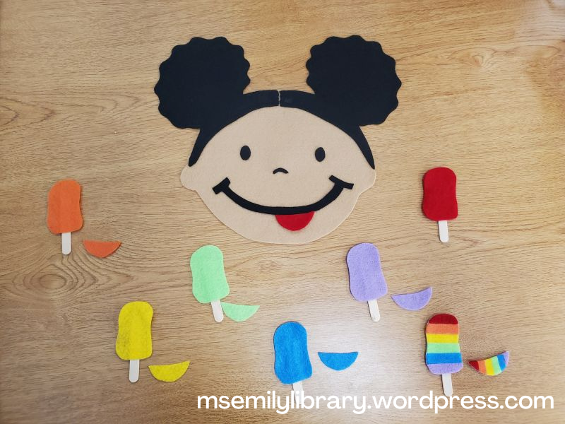photo of popsicle girl flannelboard set. includes a dark skinned girl with afro puffs and a large smile on her face, as well as 7 popsicles and a coordinating color tongue: orange, yellow, green, blue, purple, red, and rainbow.