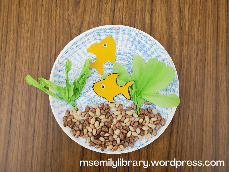 photo of fishbowl craft - paper plate colored blue, with multicolored dried beans glued to the bottom, tissue paper aquatic plants, and two goldfish with googly eyes.