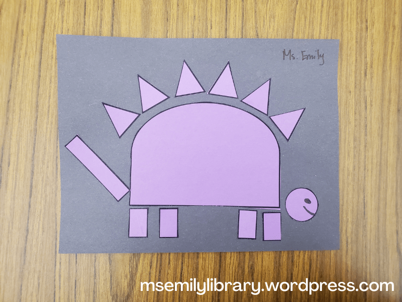photo of shape-o-saurus craft - purple shapes on a black paper make a stegosaurus.  triangles, half-circles, circles, and rectangles make up the body.