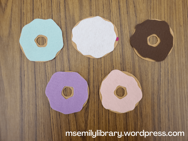 Flannel showing 4 traditional doughnuts (with light green, lavender, pink, and brown icing) and one jelly-filled doughnut with white icing and a small pink dot of jelly at one end.