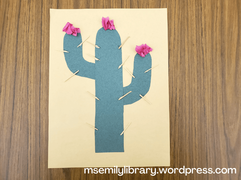 Craft of a saguaro cactus, with one main trunk and two arms, glued on a sandy colored paper.  Toothpicks are glued on as spines, and pink tissue paper flowers are glued to the top of the trunk and arms.