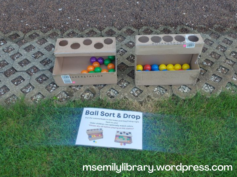Ball sort and drop station: two cardboard book boxes hold plastic "ball pit" balls.  Both have holes cut into the top with an open space below for the balls to be retrieved. 