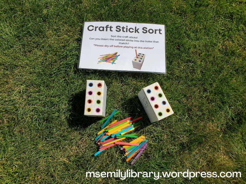 Craft stick sort station with small cardboard boxes with eight small holes in each, ringed in color, and a pile of colored craft sticks in front of them