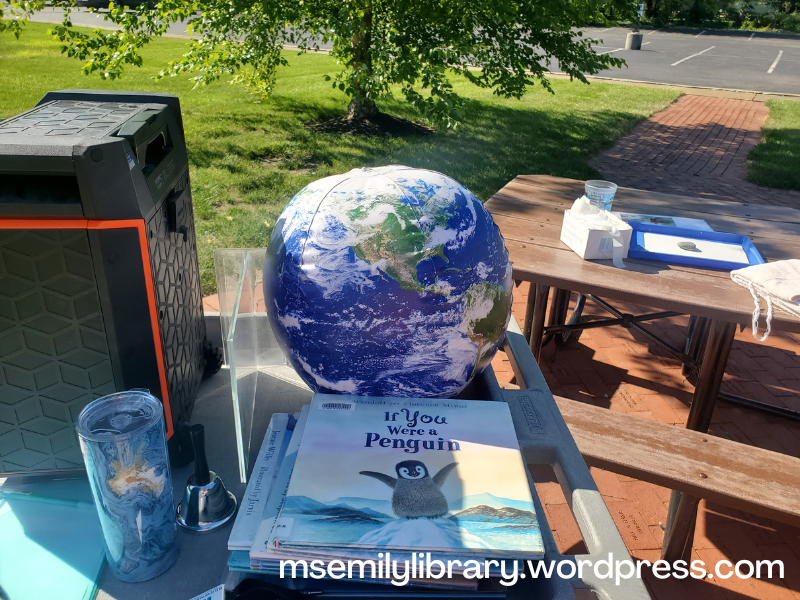 view of my storytime cart, with the speaker, my water, a stack of books (If You Were a Penguin on top) and an inflatable globe showing NASA images of the earth (land, sea, and clouds)
