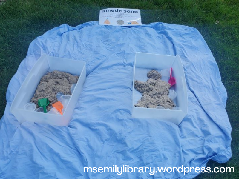 Kinetic sand station outside - two bins set on a blue sheet, with small plastic containers, plastic shovels, and sand in them