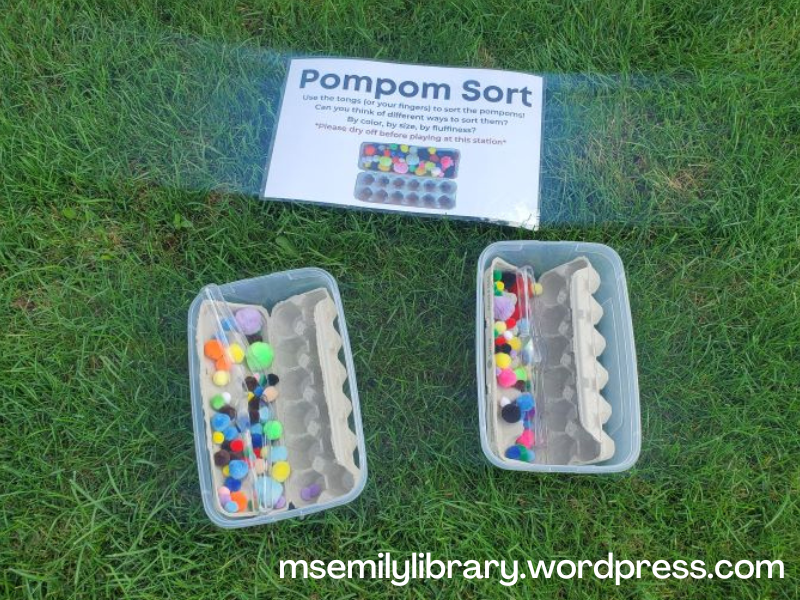 Pompom Sorting station: two shoebox sized plastic bins hold two natural cardboard egg cartons with various shaped and colored pompoms and plastic tongs.
