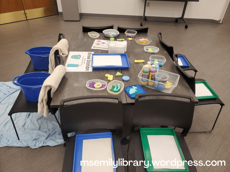 sponge painting and rinse station in the indoor setting, with painting supplies set on a square table and two chairs facing outward on each side.  Six chairs hold a messy tray and paper on them, and two hold buckets of water and towels for rinsing.