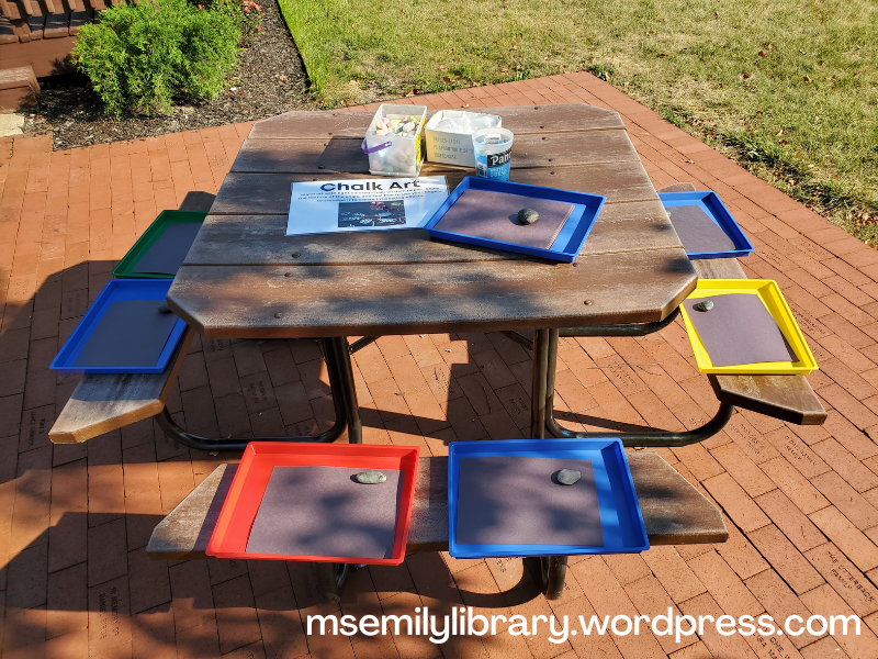 Art station on a picnic table, with messy trays set on the bench and art supplies on the table.  Includes black paper and sidewalk chalk, as well as a sign explaining the station.