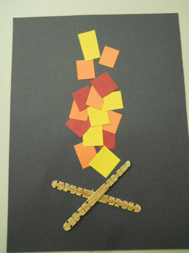 Photo of craft with red, orange, and yellow squares of construction paper glued to a black piece of construction paper, with two craft sticks glued crisscross underneath