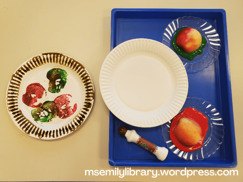 Photo of paper plate apple pie craft, with the tray of supplies to the side, including blank paper plates, red and green paint with a halved apple cut side down in the paint, and a brown dot marker.