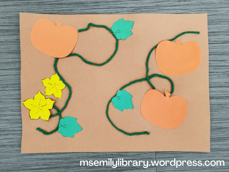 Craft showing paper orange pumpkins, yellow flowers, green leaves and green yarn representing a pumpkin vine, pasted to a brown sheet of construction paper.