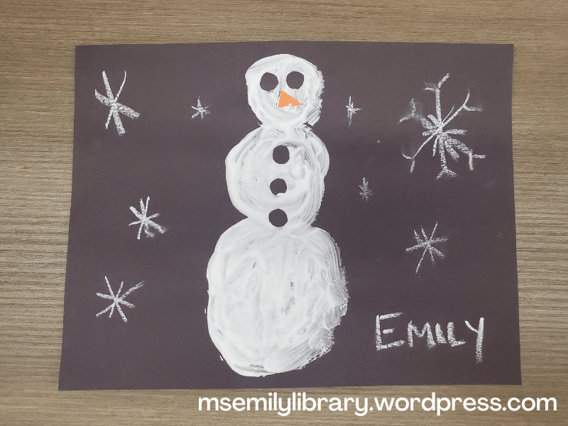 craft showing snowflakes drawn on black construction paper with white chalk, and a snowman made of white puffy paint with two black paper eyes, three black paper buttons, and a orange paper nose.