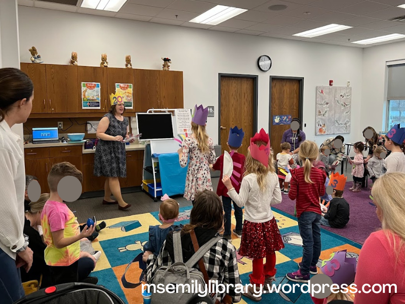 Photo from storytime, showing Emily in a yellow crown holding a noisemaker, with kids and grownups following along.