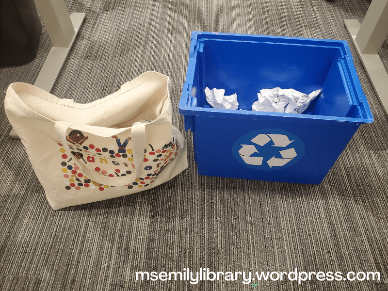 a canvas tote bag sits next to a blue recycling bin with a white recycling symbol visible on the side. Crumpled up white paper can be seen inside both.