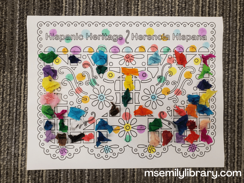 Craft of a papel picado banner, saying "Hispanic Heritage/Herencia Hispana" at the top, and showing two birds flying, among decorative flower, square, and lacy shapes. It's decorated with colored dots and multicolored scrunched up tissue paper squares.