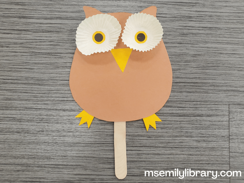 Owl puppet made of brown construction paper with eyes made from white cupcake liners with smaller yellow and black pupils glued to the middle, with a triangular beak and three toed feet, glued to a jumbo craft stick.