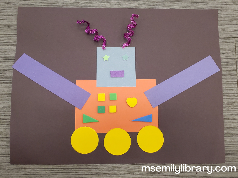 shape robot glued to a piece of black construction paper, with a long orange rectangular body, two blue arms, a light blue square head, purple metallic squiggly antennae, three yellow wheels, and variously colored buttons, eyes, and mouth.