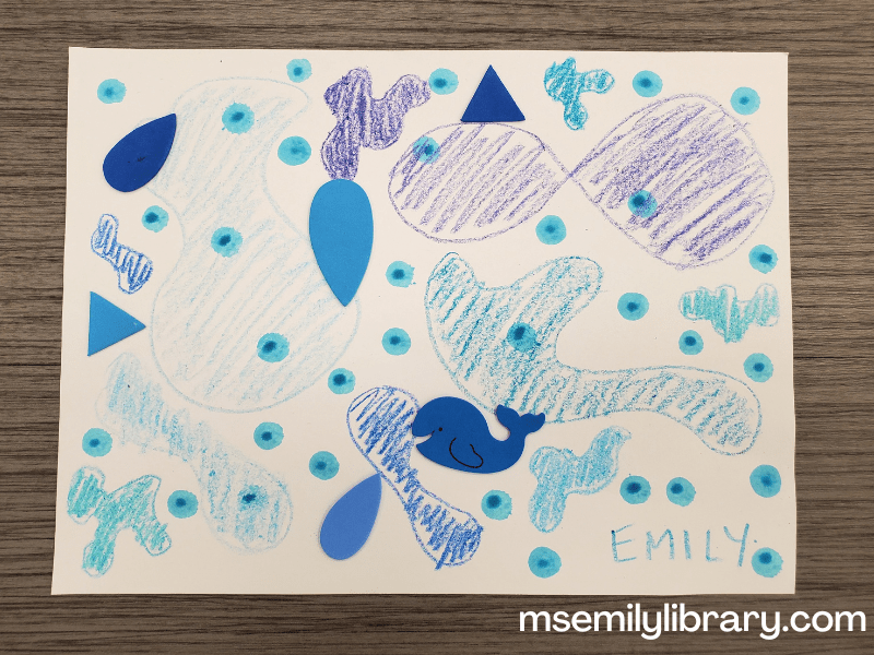 picture of craft: blue abstract blob shapes are drawn in various shades of blue crayon. Blue dots from a dot marker are all over the page, and different shaped foam stickers (teardrops, triangles, and a whale) are also glued on.