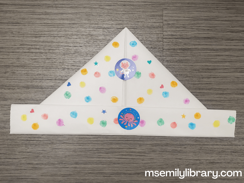 an origami hat decorated with multicolored dots from dot markers, plus heart and star stickers and two round stickers of animals in space.