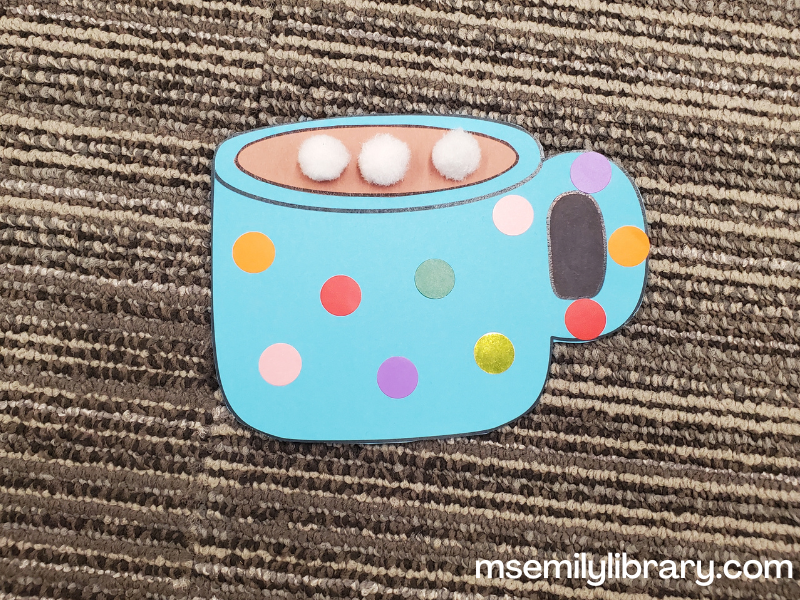 blue hot chocolate mug with an oval of brown construction paper on top, decorated with colorful dot stickers. Three white pompoms make the marshmallows on top.