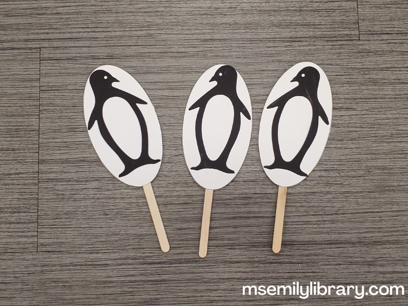 Three paper penguin puppets - a white oval is attached to a craft stick, and a black die-cut penguin is glued to the white oval.