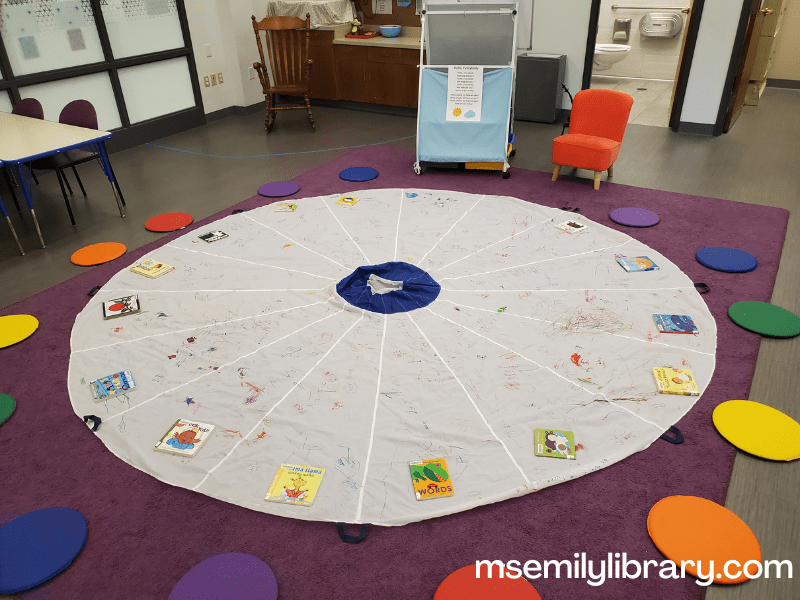 parachute set up showing a white parachute on a purple rug, with rainbow colored circular mats arranged around the edge and board books in front of every mat. At the head of the room is a small pink upholstered chair and an easel with rhyme sheets taped to it.