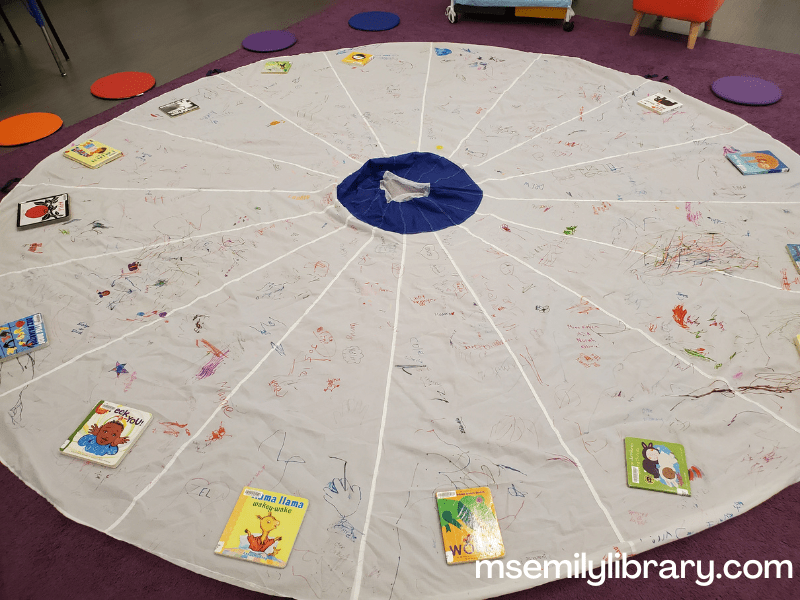 closer photo of white parachute, showing colorful signatures, drawings, scribbles, hand prints, etc. board books are arranged at the end of each wedge and circular mats are on the floor around the edge.