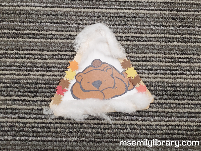 Bear in a cave craft consisting of a printed picture of a sleeping bear glued to a triangle of wooden craft sticks. fall-colored leaves are glued to the sides and cotton is glued to the top and bottom to simulate snow.