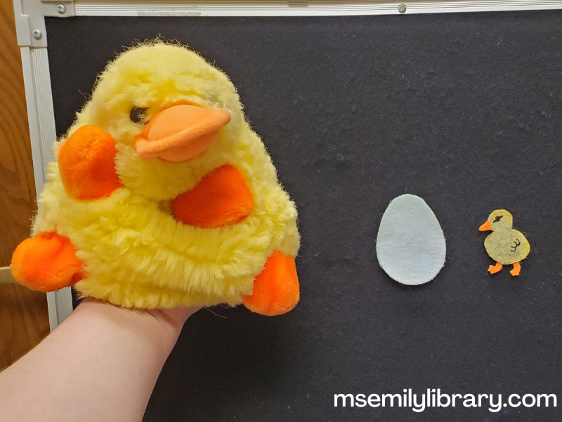 Duckling flannel and puppet - a fat yellow duck puppet poses in front of the flannelboard, which shows a blue-green egg and duckling flannel pieces.