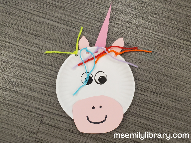 paper plate unicorn craft with a small paper plate decorated with a pink horn, ears, and snout, sticker eyes, and a multi colored yarn mane.