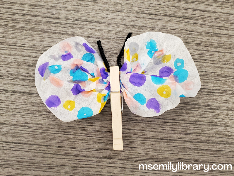 coffee filter butterfly craft, showing a white coffee filter pinched in the middle to create two wings. It's decorated with blue yellow, and purple dots, and secured with a wooden clothespin.