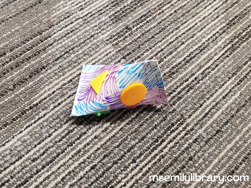 photo of noisemaker craft - a toilet paper tube stapled at both ends (perpendicular angles) and decorated with markers and foam stickers.