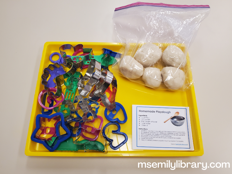 messy tray with a number of plastic and metal cookie cutters, a gallon size zip top bag with five balls of neutral colored homemade playdough, and quarter sheet paper with the recipe for homemade play dough.