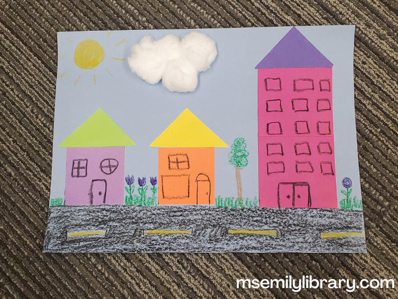 photo of craft, showing two small houses and one tall apartment building, with a road, landscaping, a sun, and cotton ball sky. The buildings are made with construction paper shapes, and the rest is crayon.