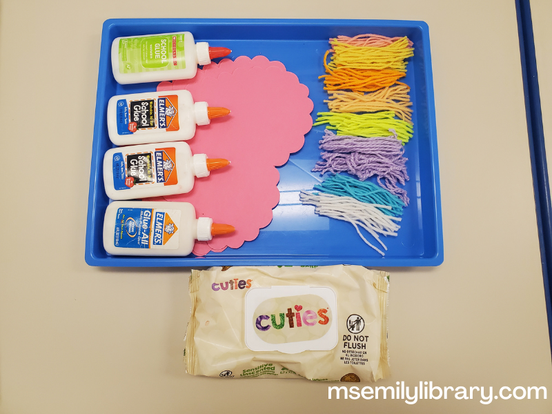photo of craft setup, showing piles of different colored yarn, pink paper hearts, and liquid glue on a messy tray, with a package of baby wipes on the table next to it.