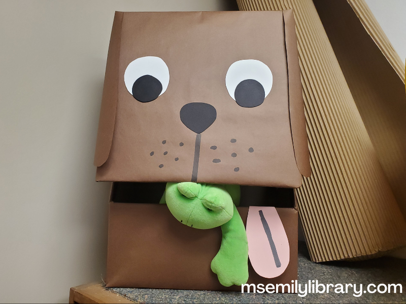 bark george puppet, made from a box covered in brown paper with eyes, muzzle, ears, and tongue added. The mouth is where the box opens on a hinge. Shown in this picture with a frog puppet halfway out of the mouth.