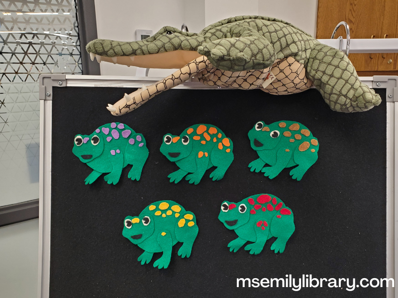 flannel of 5 green frogs, each with different colored speckles. Above the flannelboard, an alligator puppet