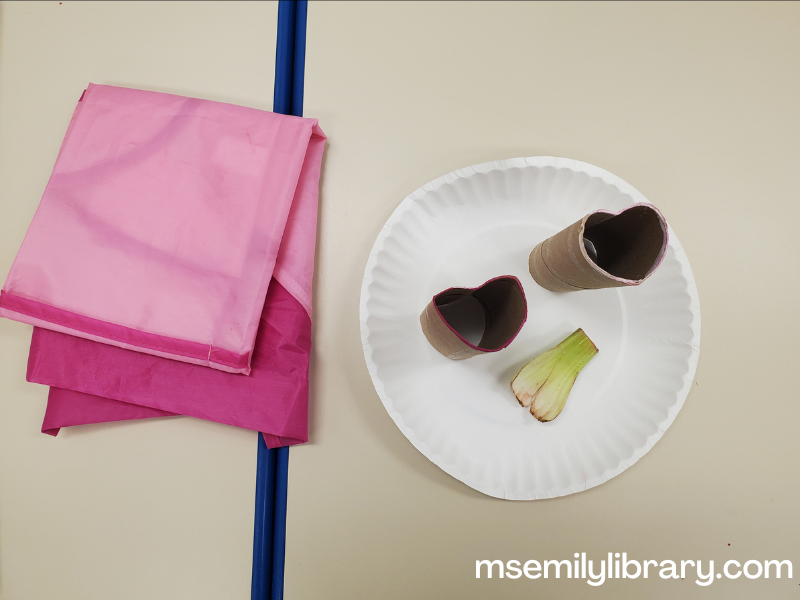 toilet paper tubes bent into heart shapes, with a piece of celery on a paper plate. also pictured is a pink art smock folded up.