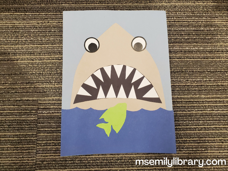 Shark craft showing a large shark face looking down at the water in front o fit where a little green fish is swimming. There are lots of sharp white teeth!