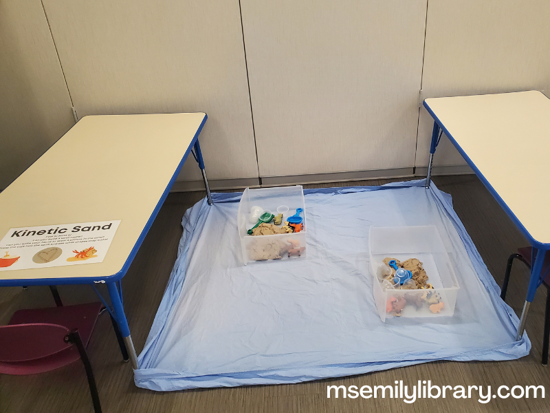 kinetic sand station, showing a fitted sheet held down by two tables, and two clear bins with sand and plastic farm animal figurines