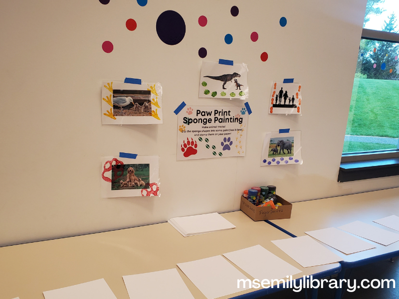 animal paw print art station showing a table with blank paper and paint, and on the wall is a sign for the station and examples of art