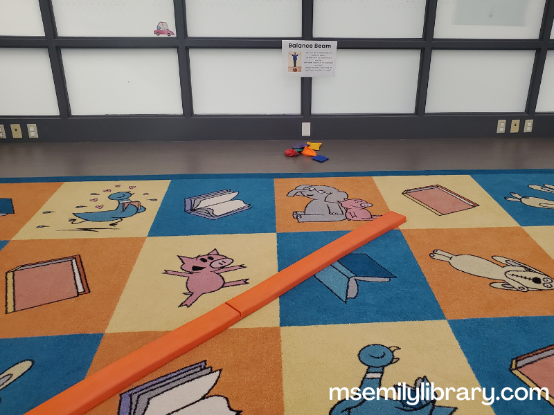 balance beam station, with an orange rubber balance beam on a rug, with a sign explaining what to do. a pile of bean bags is nearby.