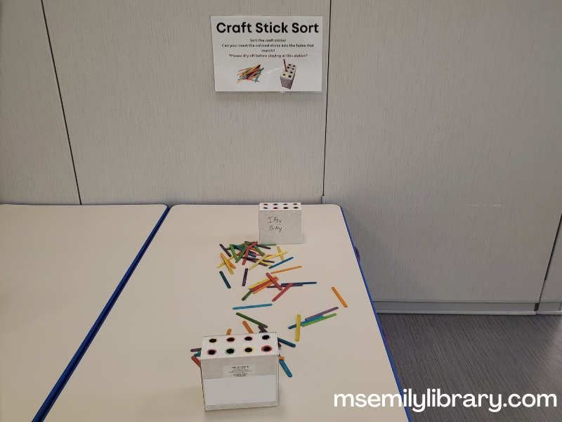 Craft stick sort station, showing colored craft sticks strewn on a table with two small boxes with color coded holes.