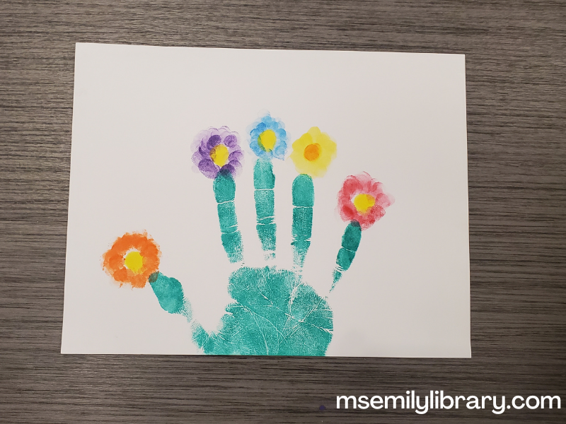 Craft showing a stamped hand in green. Each finger tip has a flower stamped in different colors.