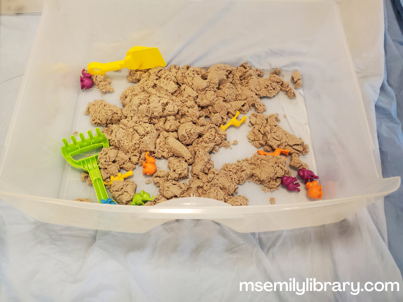 closeup of a kinetic sand bin, showing the bin, some plastic shovels, and plastic animals
