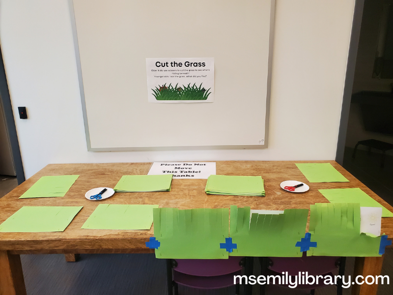 cut the grass station, showing green construction paper sheets with 1 inch strips cut halfway down to make grass. Underneath are papers with various things that may be found in grass: flowers, apples, bugs, etc.