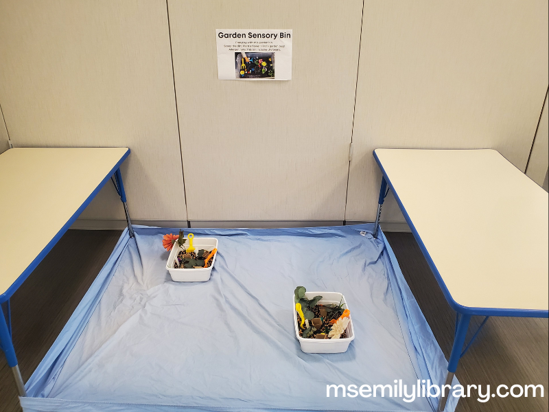 garden sensory bin, showing two bins on a fitted sheet with black beans and various gardening toys like shovels, plastic bugs, fake flowers, plant labels, and small seed starter containers